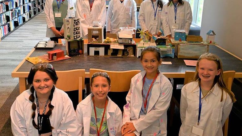 A group of young students in lab coats posing in two rows in front of and behind a table.